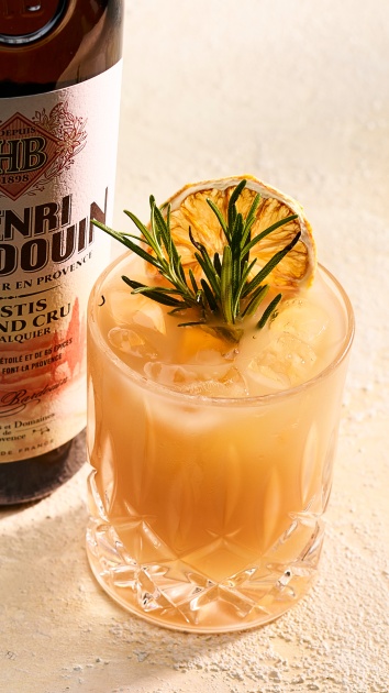 Ein Cocktail in einem Kristallglas, garniert mit Rosmarin und einer Zitronenscheibe neben einer Flasche.