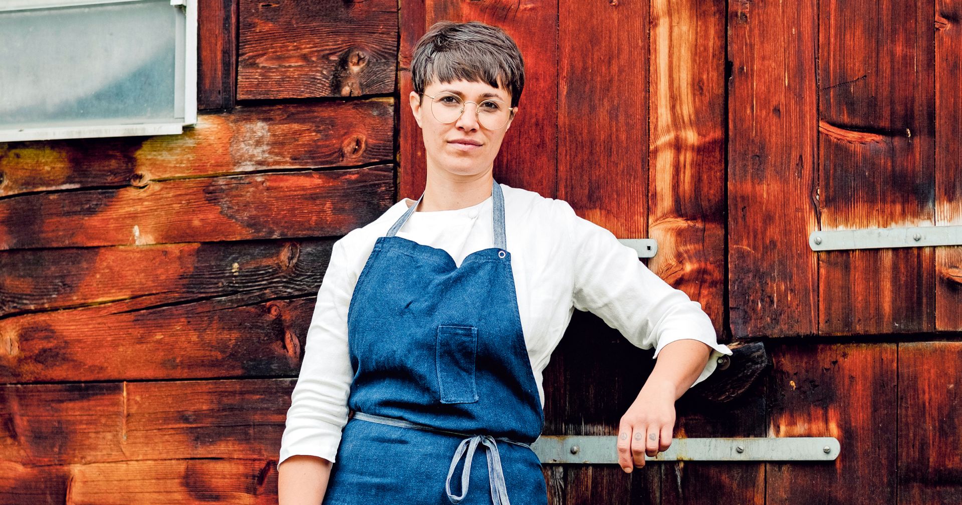 Foto einer Frau mit Kurzhaarschnitt und blauer Schürze vor einer rustikalen Holzwand.