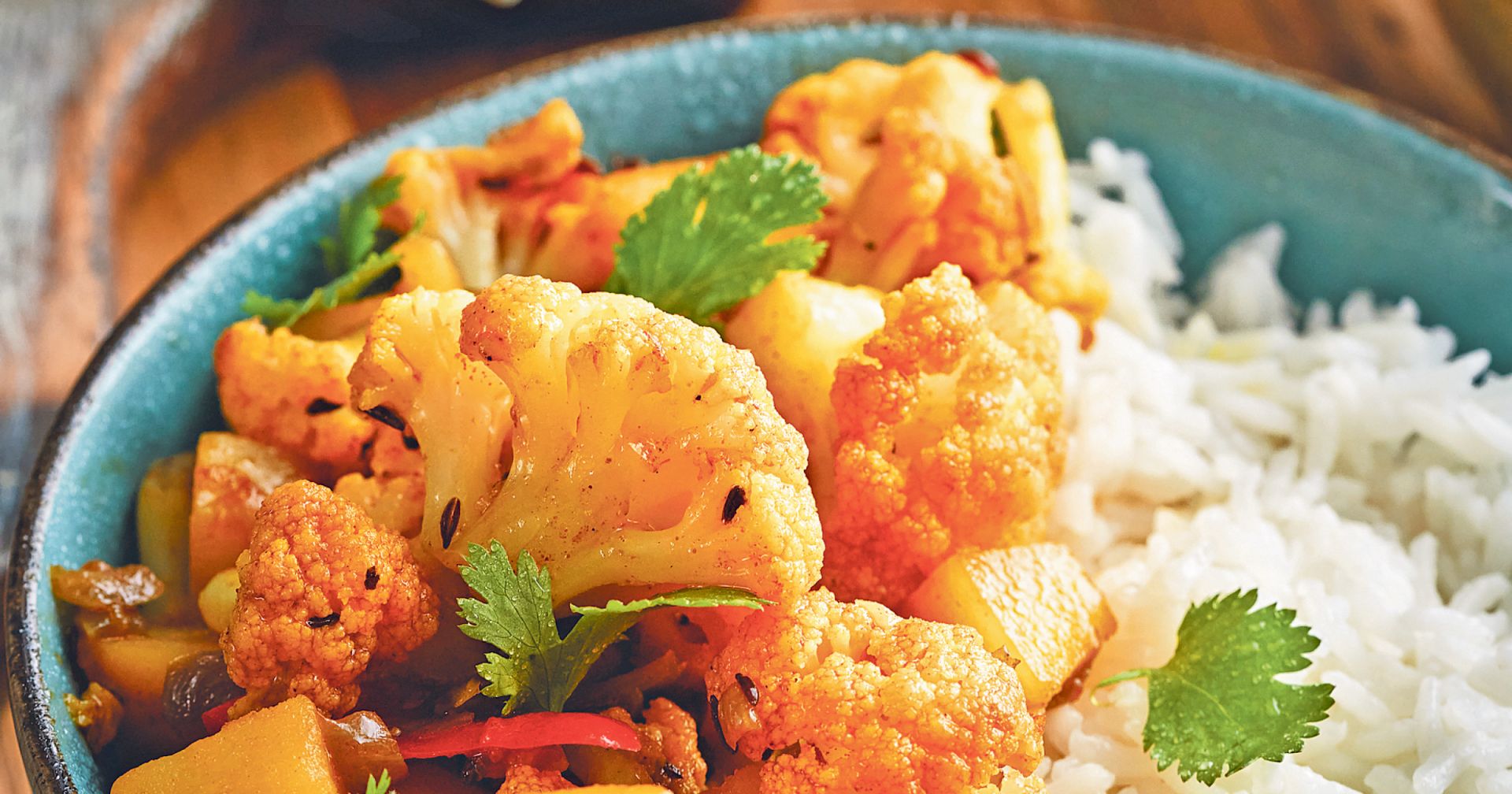 Blaues Schälchen mit würzigem Blumenkohl-Curry und weißem Reis, garniert mit frischem Koriander.