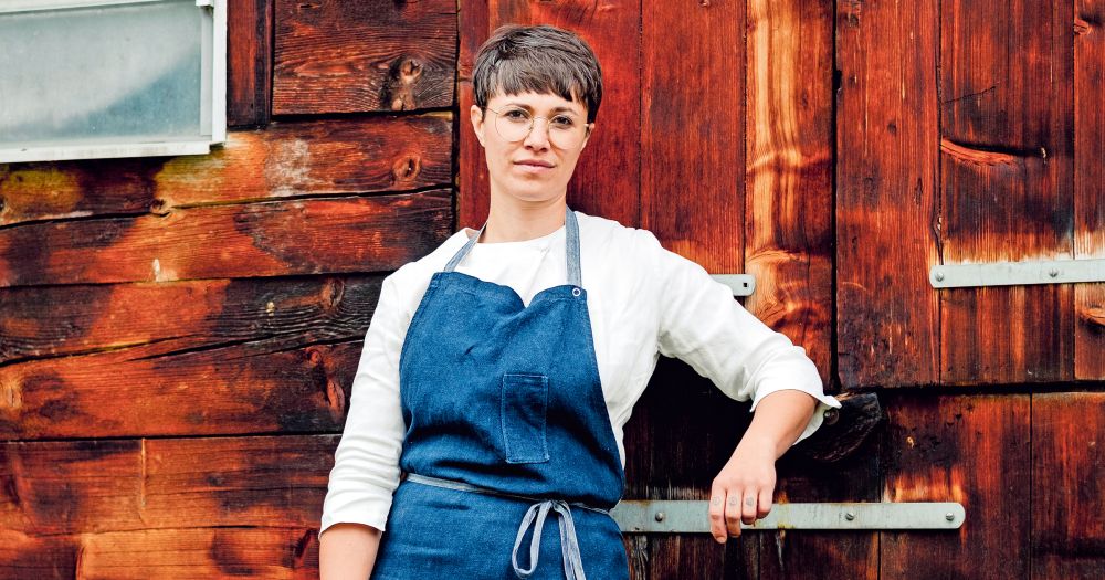 Foto einer Frau mit Kurzhaarschnitt und blauer Schürze vor einer rustikalen Holzwand.
