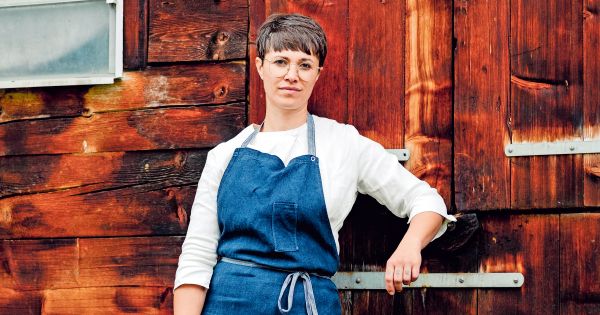 Foto einer Frau mit Kurzhaarschnitt und blauer Schürze vor einer rustikalen Holzwand.