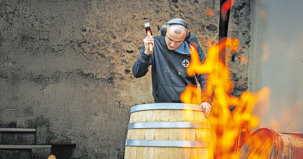 Ein Küfer bearbeitet mit einem Hammer ein Holzfass neben einem offenen Feuer.
