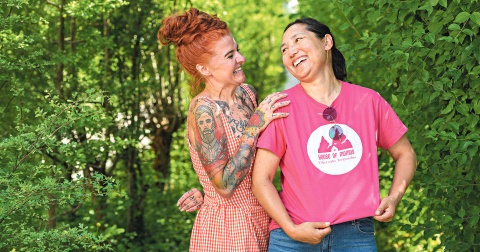 Zwei lachende Frauen stehen eng beieinander vor einer grünen Hecke im Freien.