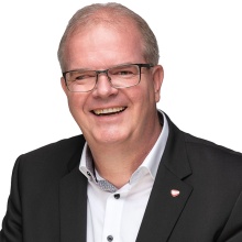 Porträtfoto eines lächelnden Mannes mit Brille, weißem Hemd und schwarzem Sakko.