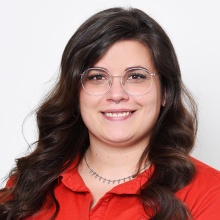 Porträt einer lächelnden Frau mit braunem Haar und Brille vor weißem Hintergrund.