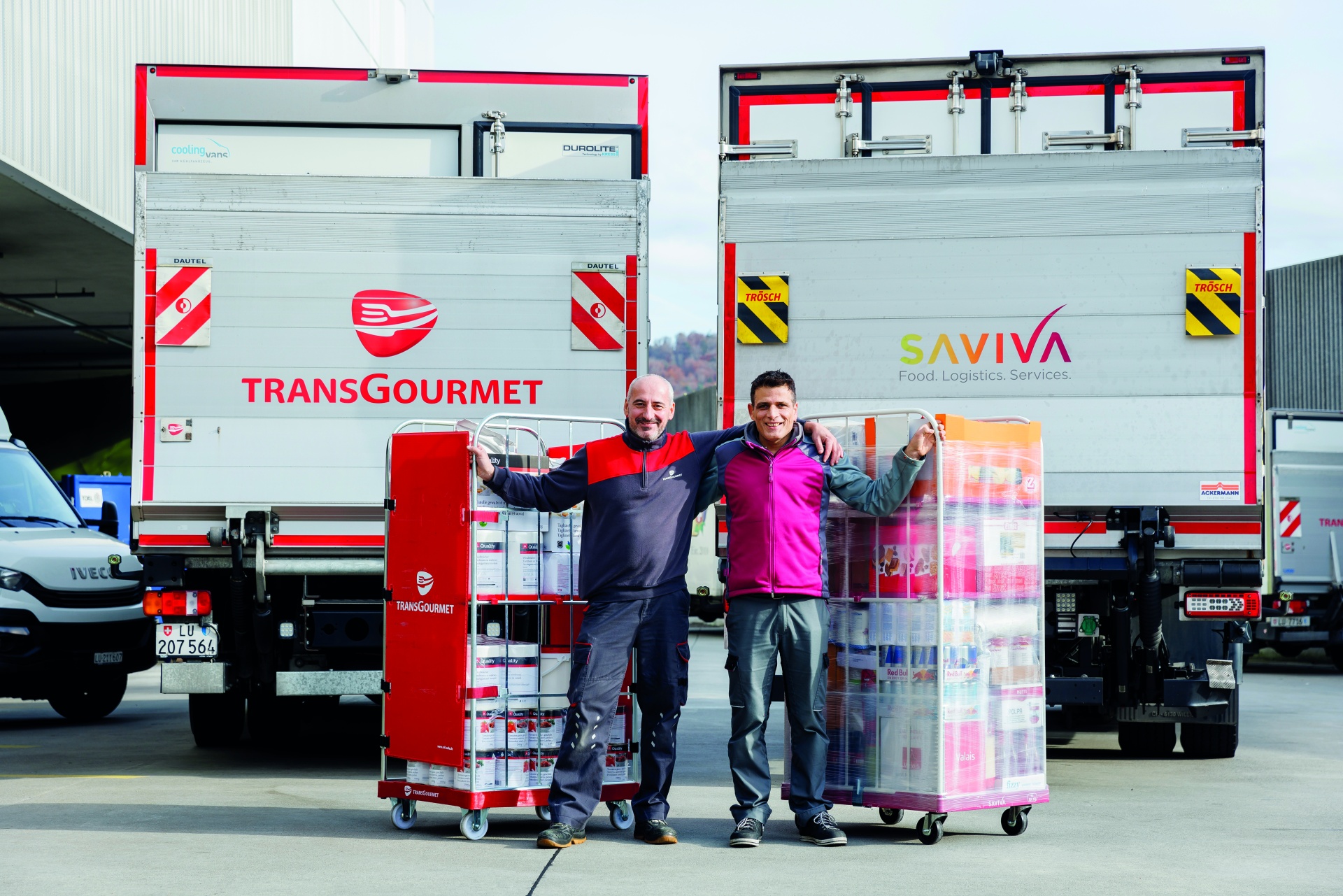 Die beiden Lastwagen von Transgourmet und Saviva stehen nebeneinander. Transgourmet und Saviva Mitarbeiter vereint.